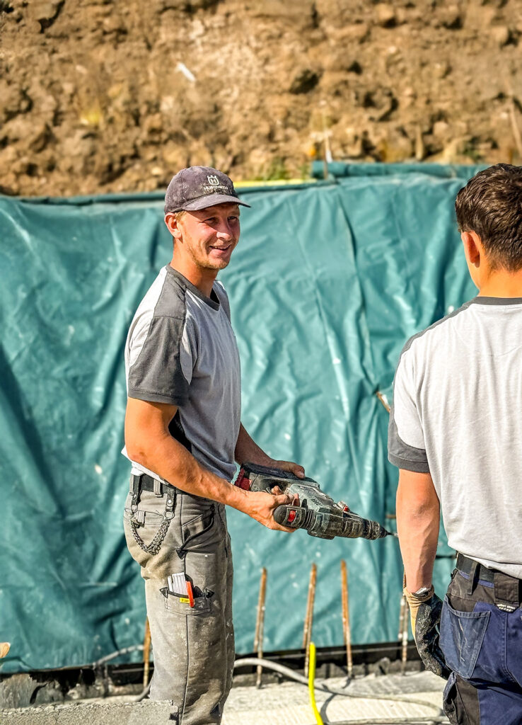 Zwei Personen in Arbeitskleidung stehen auf einer Baustelle, eine Person hält ein handgeführtes Elektrowerkzeug