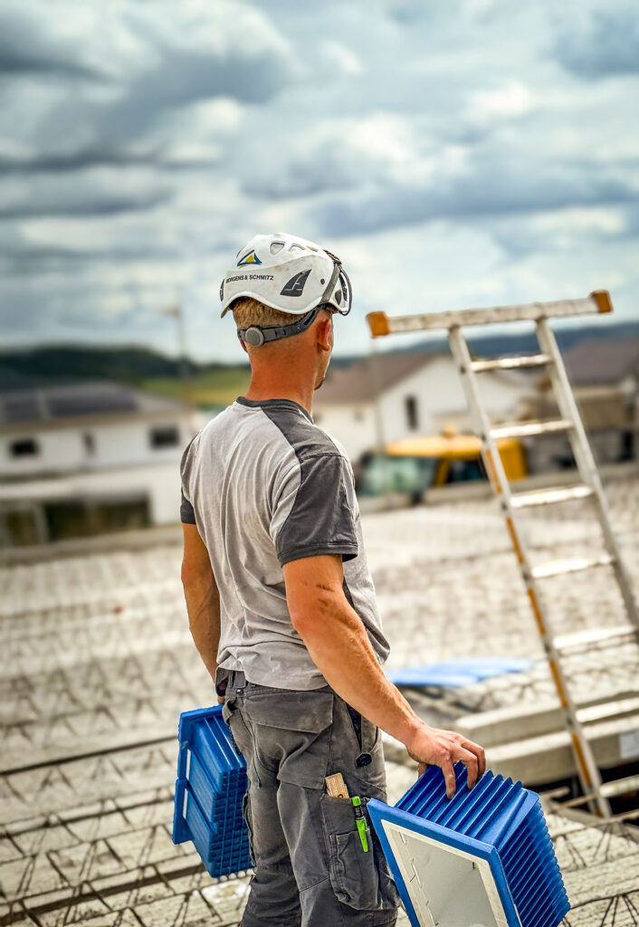 Person in Arbeitskleidung und Schutzhelm trägt auf einer Baustelle Bauteile über eine Betondecke