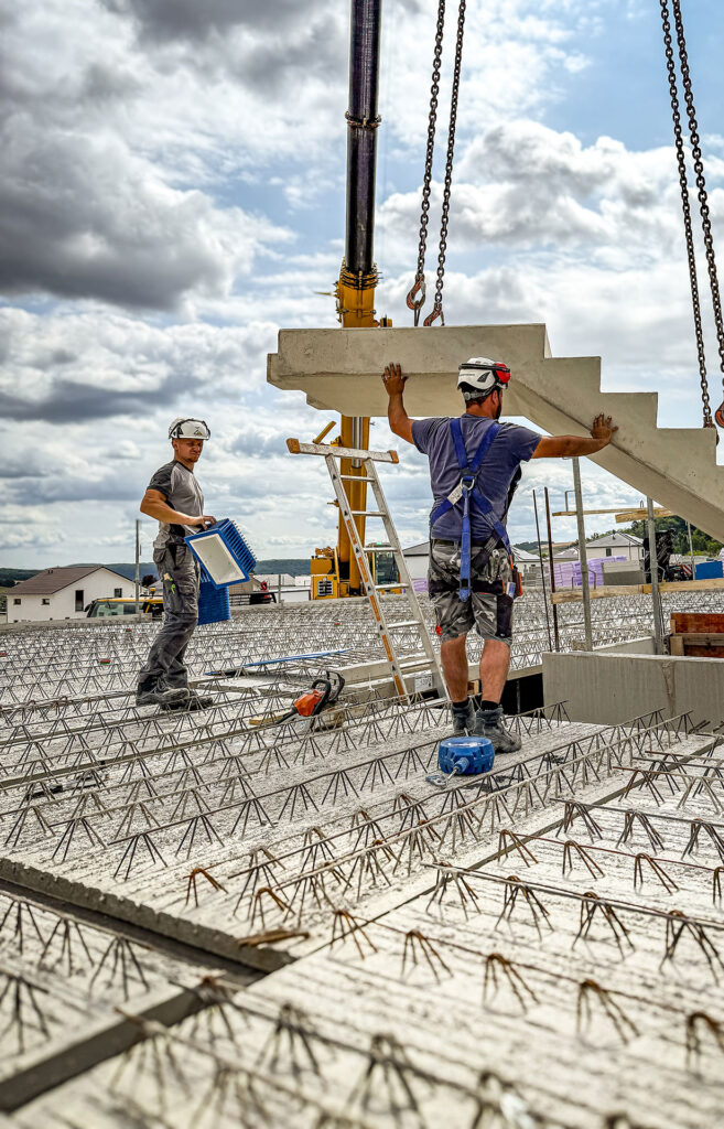 Zwei Personen in Arbeitskleidung positionieren auf einer Baustelle ein Betonelement mit Hilfe eines Krans
