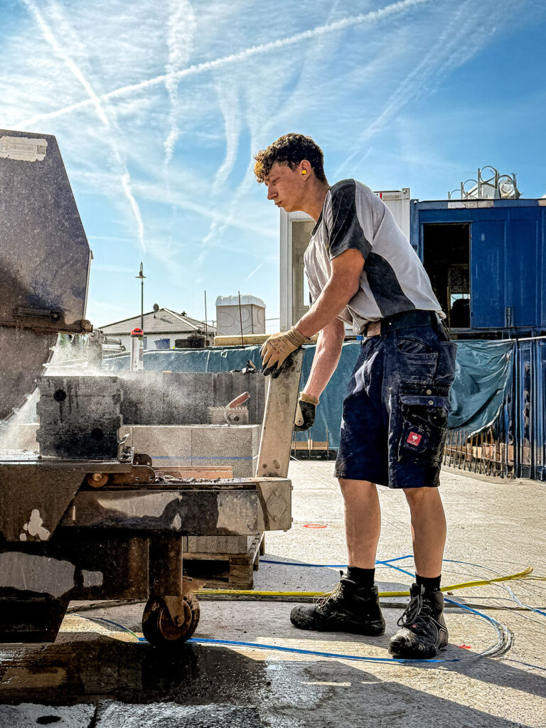 Person in Arbeitskleidung bedient auf einer Baustelle eine Steinsäge und bearbeitet einen Baustoff